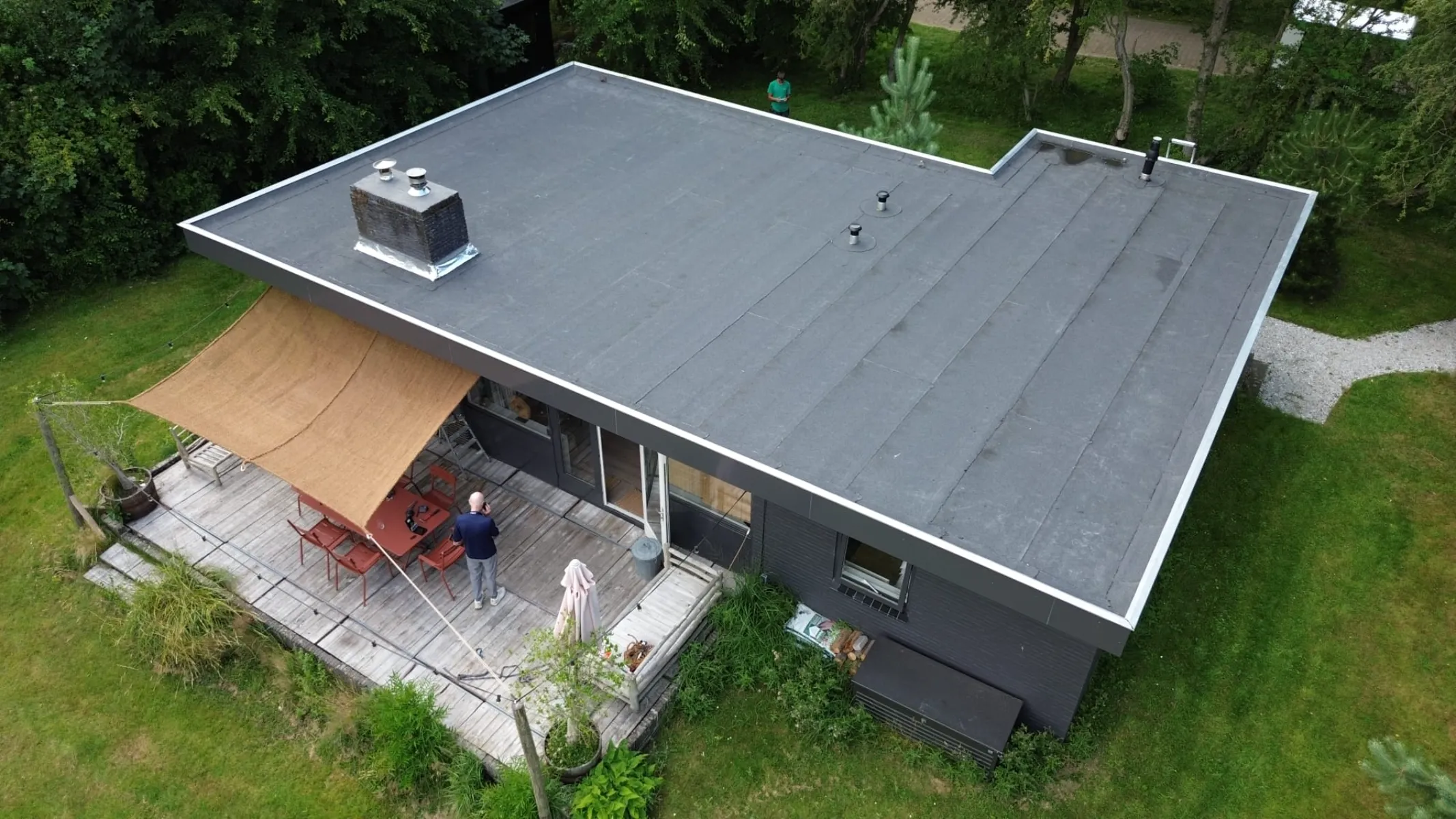 Luchtfoto van platte daken op woning met houten terras, schaduwdoek en omliggende begroeiing