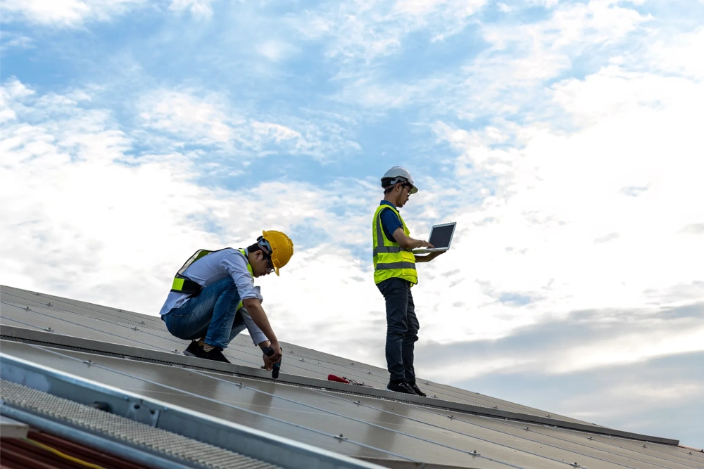Twee dakwerkers met helm en veiligheidsvest plaatsen panelen op een schuin metalen dak met gereedschap en laptop